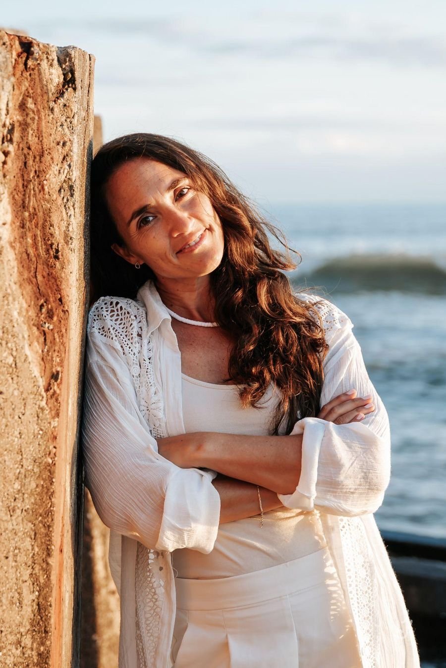 Paula Florez sonriendo, apoyada en un poste de madera frente al mar, con los brazos cruzados